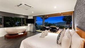 A modern bedroom with a large bed, a freestanding bathtub, a TV, and a balcony with a view of the landscape.