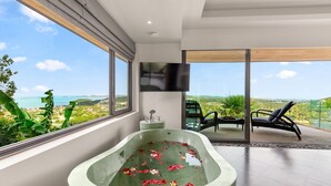 A modern bathroom with a freestanding bathtub filled with water and rose petals, large windows offering a scenic view of the sea and greenery, and a flat-screen TV mounted on the wall.