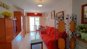 Ein Wohnzimmer mit einem roten Sofa, einem Glastisch und einer Holzkommode. Im Hintergrund befindet sich ein Essbereich mit Tisch und Stühlen.