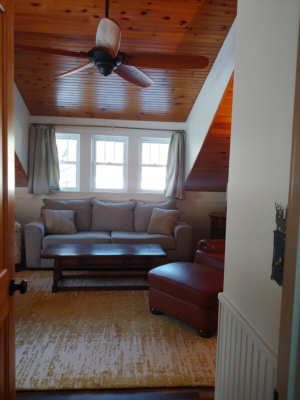 Ein gemütliches Schlafzimmer im Loft-Stil mit Holzbalkendecke, Deckenventilator, großem Fenster mit Vorhängen, einer Couch, einem kleinen Tisch und einem Koffer.