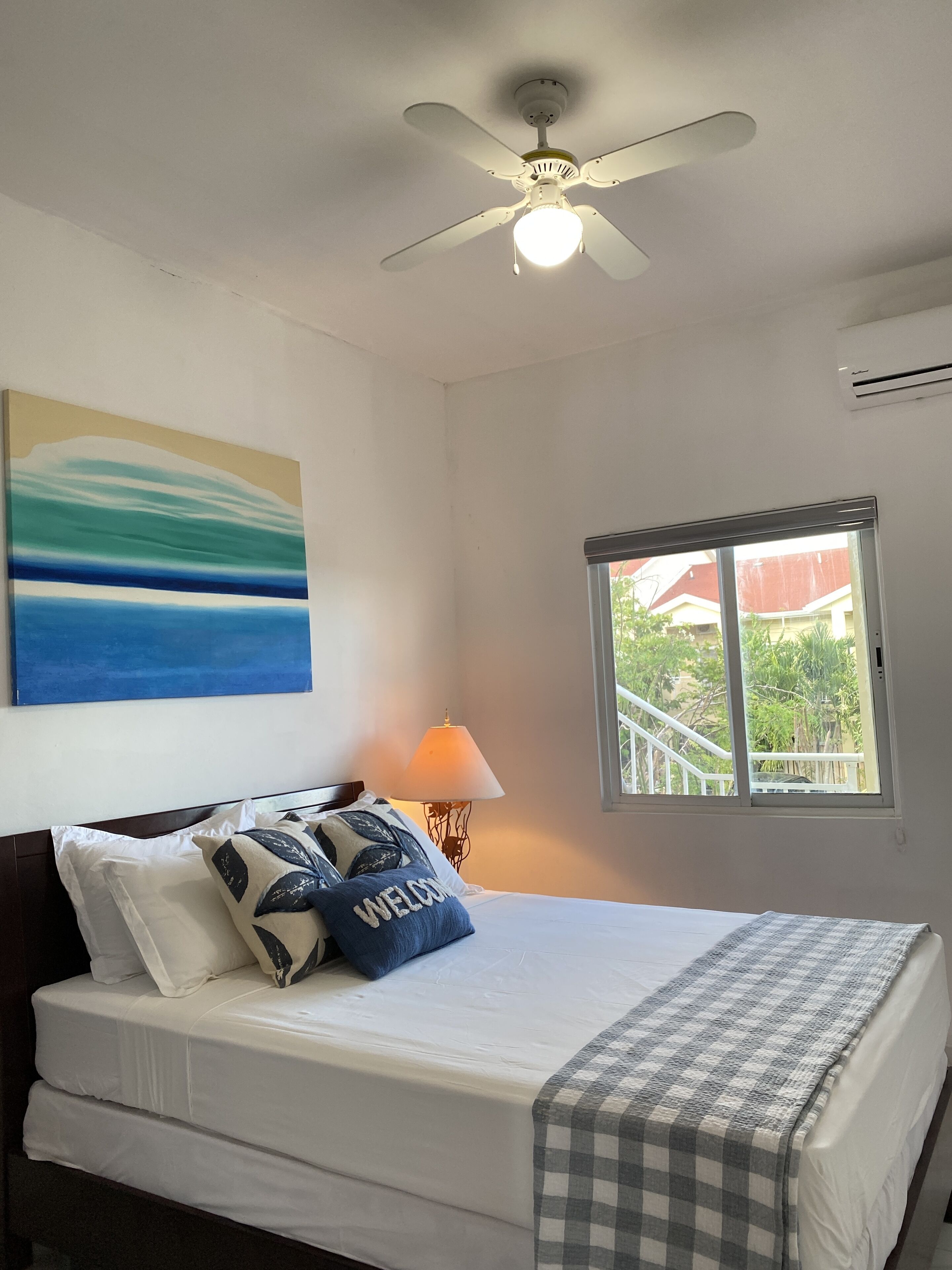 Un dormitorio con cama, ventilador de techo, ventana con vistas, un cuadro colgado en la pared y una lámpara de noche.