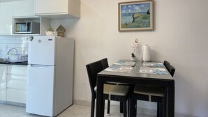 A modern kitchen with white cabinetry, a built-in microwave, and a white refrigerator. There is a dining table with a patterned tablecloth and two chairs. A painting of a seascape hangs on the wall above the table.