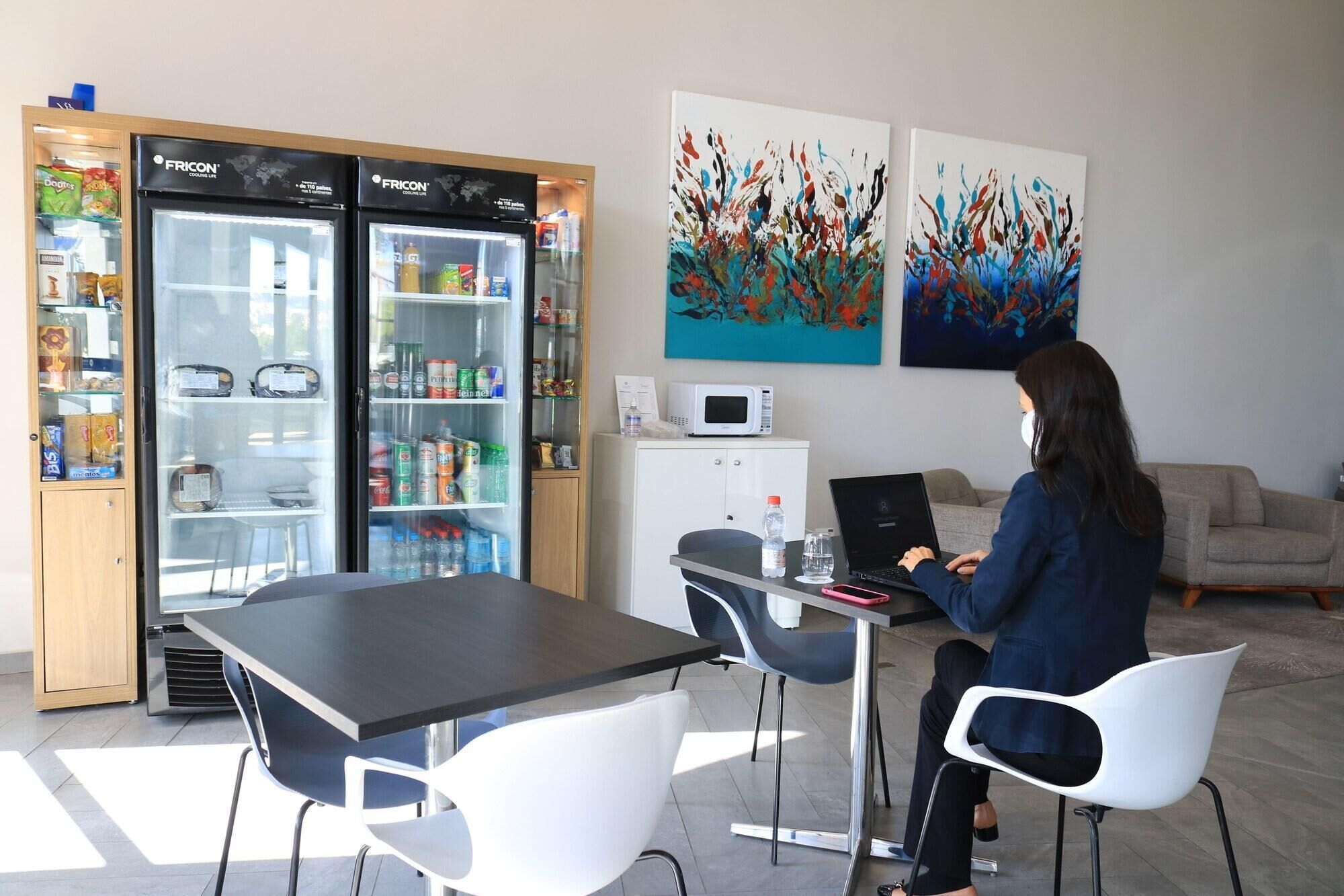 A person working at a desk with a laptop, a refrigerator with snacks, and abstract paintings on the wall.