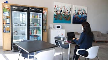 A person working at a desk with a laptop, a refrigerator with snacks, and abstract paintings on the wall.