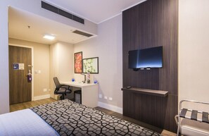 A hotel room with a bed, desk, chair, TV, and artwork on the wall.