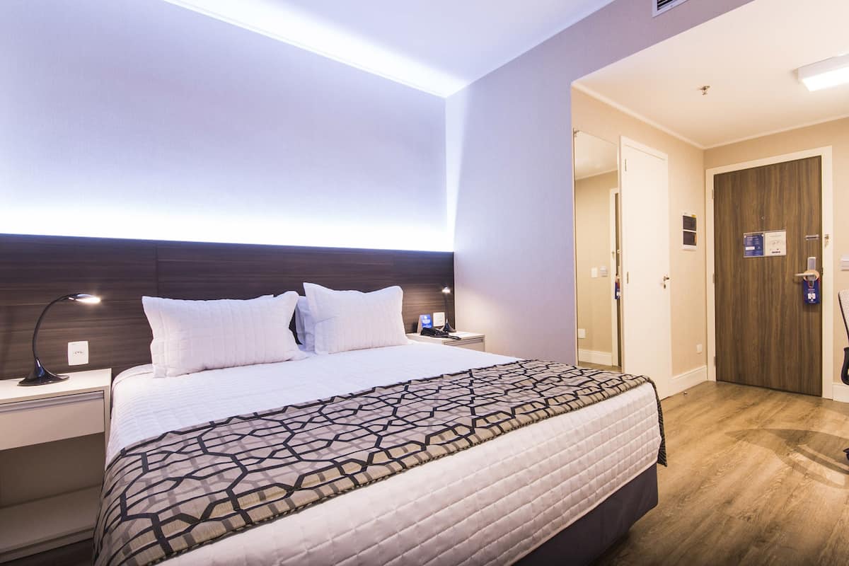 Une chambre d’hôtel comprenant un lit, des tables de chevet, une commode, un bureau, une chaise et une porte en bois.
