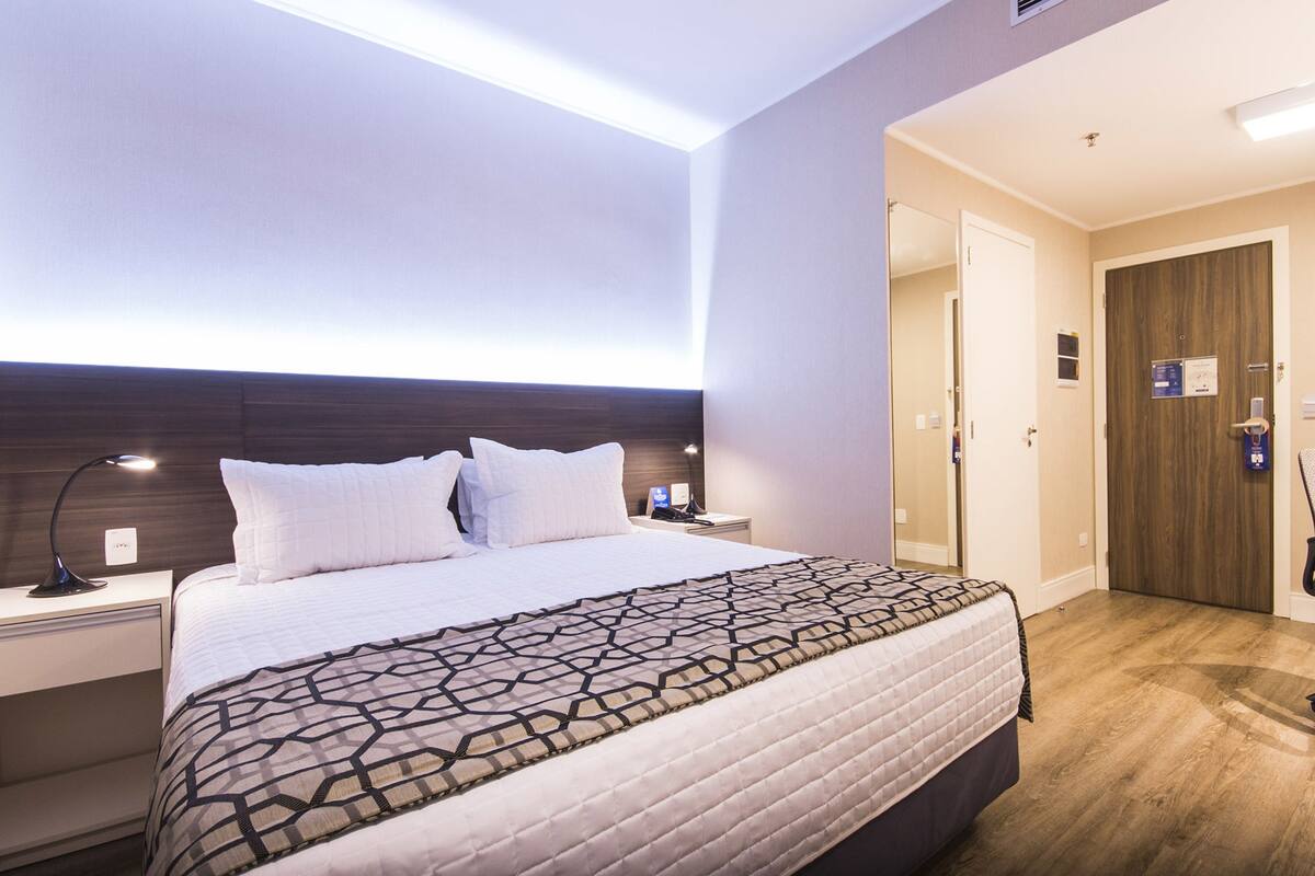 Hotellihuone, jossa on sänky, yöpöydät, yölamppu, työpöytä, tuoli ja puinen ovi.