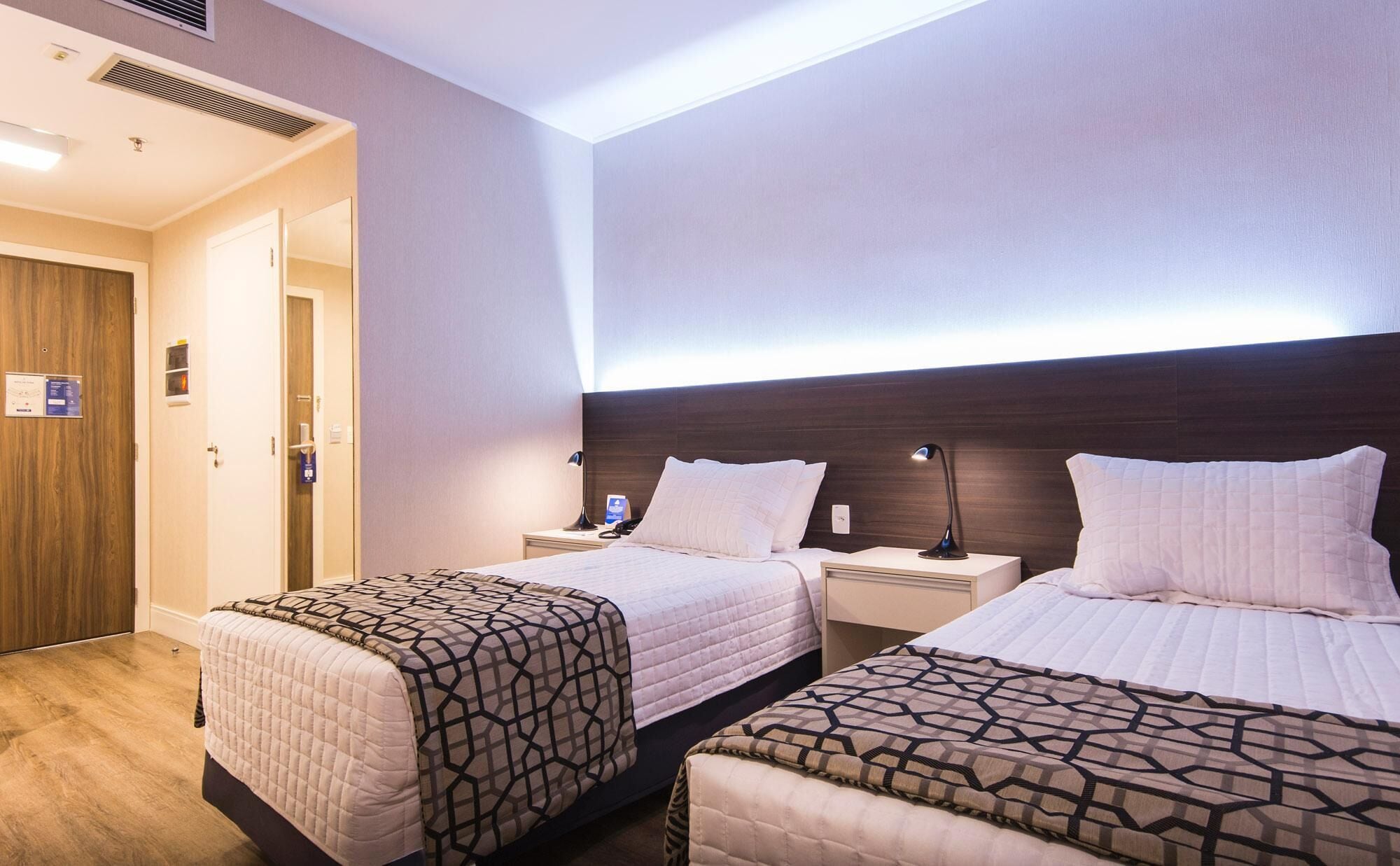 Habitación de hotel con dos camas, un armario de madera, una mesita de noche con lámpara y una rejilla de ventilación en el techo.