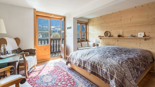 A bedroom with a large bed, a wooden headboard, a small table with chairs, a red and blue patterned rug, and a view through a glass door.