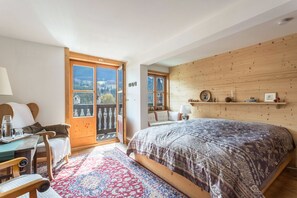 Ein Schlafzimmer mit einem großen Bett, einem hölzernen Kopfteil, einem kleinen Tisch mit Stühlen, einem Teppich mit blau-rotem Muster und Blick durch eine Glastür.