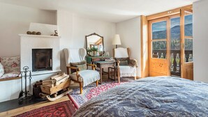 A bedroom with a bed, a fireplace, a desk, two chairs, a window with a view, and a wooden door.