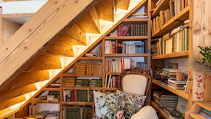 A cozy attic room with wooden bookshelves filled with books, a comfortable chair with a floral pillow, and a rustic wooden ceiling.