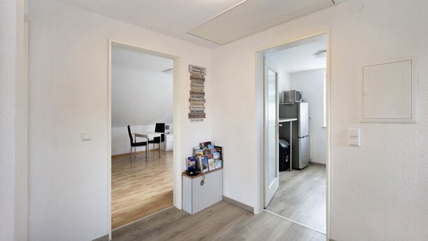 A modern interior with a wooden floor, a white wall-mounted shelf with books, and a kitchen area with a refrigerator and stove. - Holiday Apartment "Sonnenbühl" with Lake View, Private Balcony and Wi-Fi (Aulendorf)