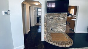 A modern living room with a stone fireplace, a flat-screen TV mounted above it, and a kitchen visible through an open doorway.