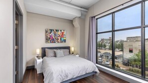 Un dormitorio moderno con una ventana grande, una cama, una mesita de noche y un cuadro abstracto colorido en la pared.