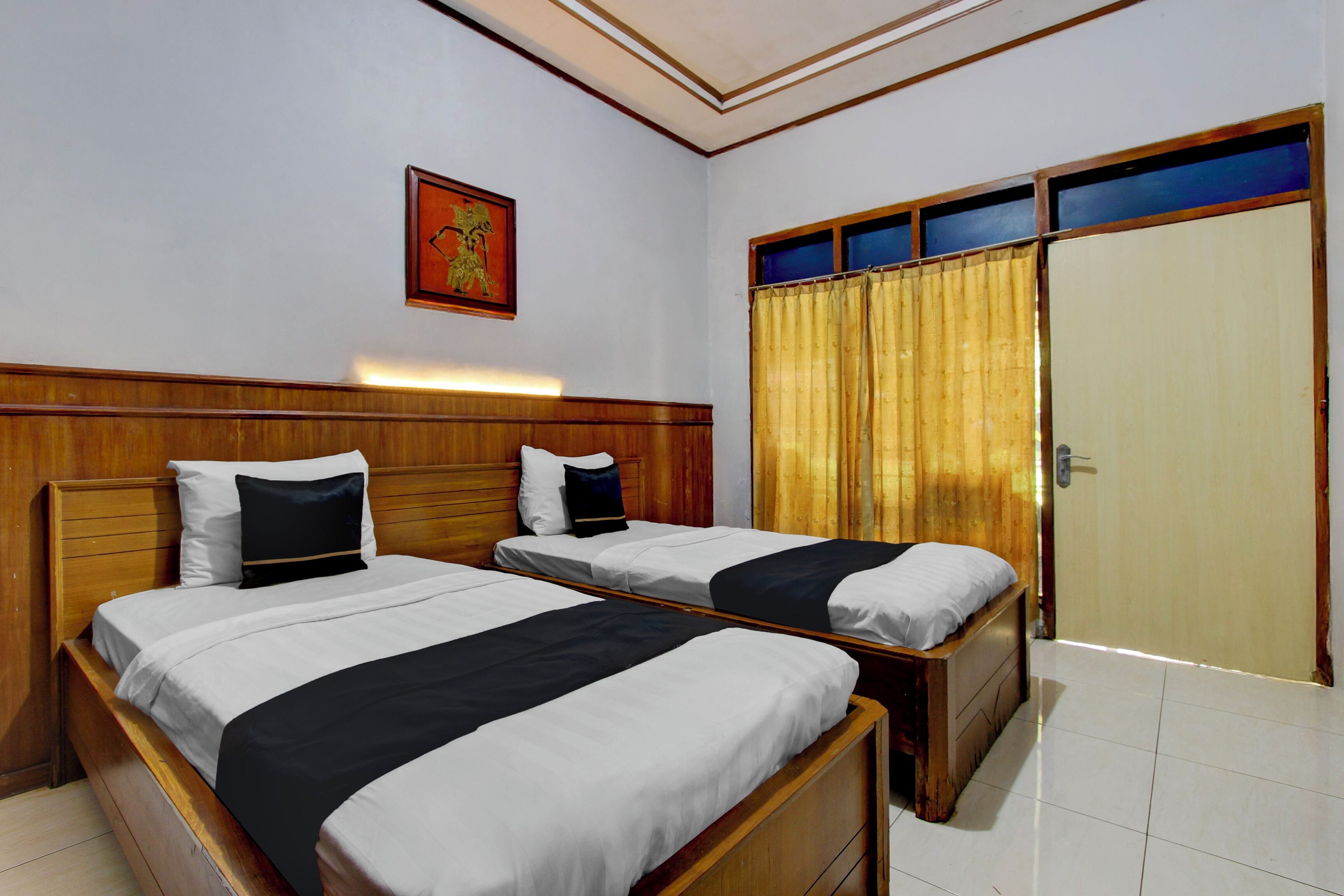 Habitación de hotel con dos camas, cabeceros de madera, un ventanal con cortinas amarillas y una puerta de madera.