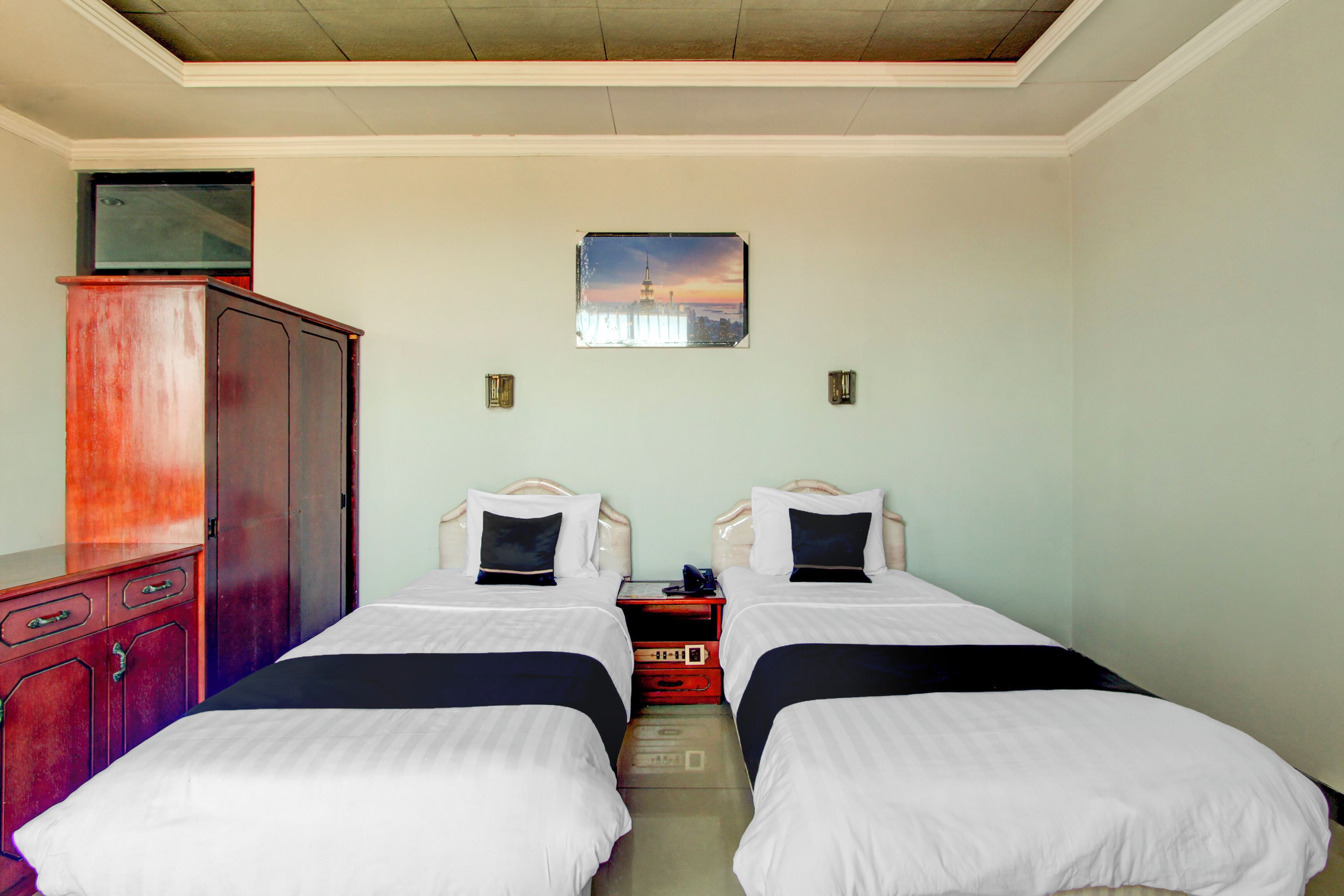Une chambre d’hôtel avec deux lits simples, une armoire en bois, une table de chevet avec un téléphone et un tableau représentant un paysage urbain accroché au mur.