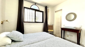 Une chambre à coucher comprenant un lit, une table de chevet, un miroir, une fenêtre avec des rideaux et un luminaire fixé au mur.