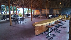 A covered outdoor seating area with a long wooden bar and multiple stools. Tables and chairs are arranged around the bar. - LAKE NYASA RESORT & CAMPSITE  (Matema)