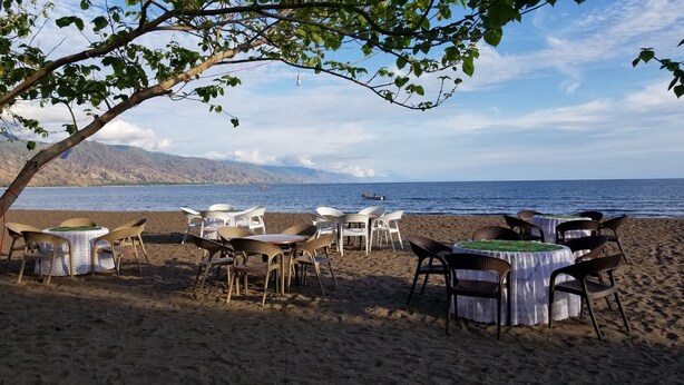 A beachside dining area with tables and chairs, overlooking a calm sea and distant mountains. - LAKE NYASA RESORT & CAMPSITE  (Matema)