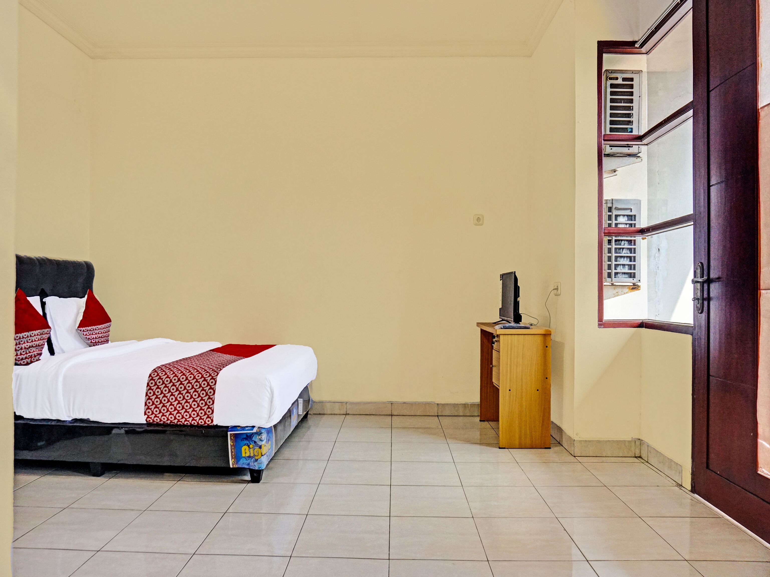 A bedroom with a bed, a small wooden nightstand, and a window with an air conditioning unit.