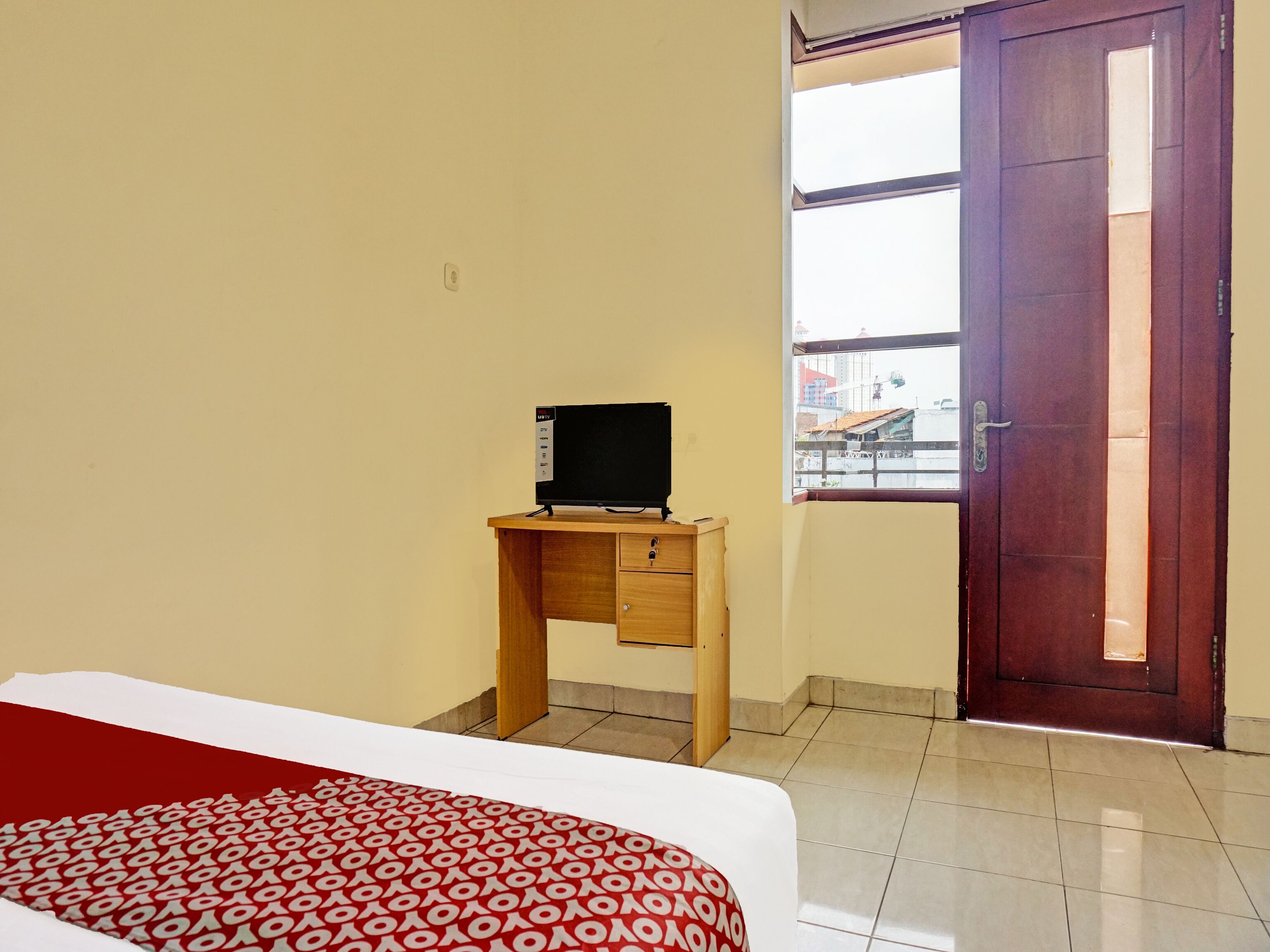A hotel room with a bed, a wooden desk with a TV, a window with a view of buildings, and a door.
