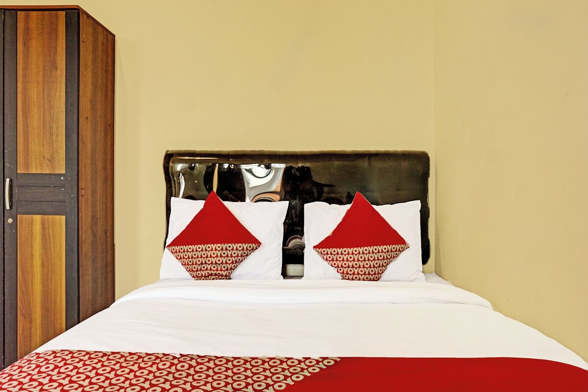 Uma cama com roupa de cama branca e almofadas vermelhas, um guarda-roupa de madeira e uma parede lisa.