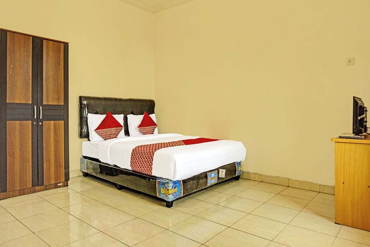 A neatly made bed with white and red bedding, a wooden wardrobe, and a small wooden desk with a television.