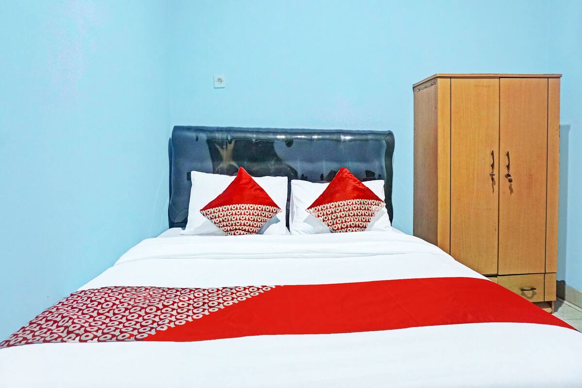 A bed with a blue headboard, two red pillows, and a white and red bedspread. There is a wooden wardrobe to the right.
