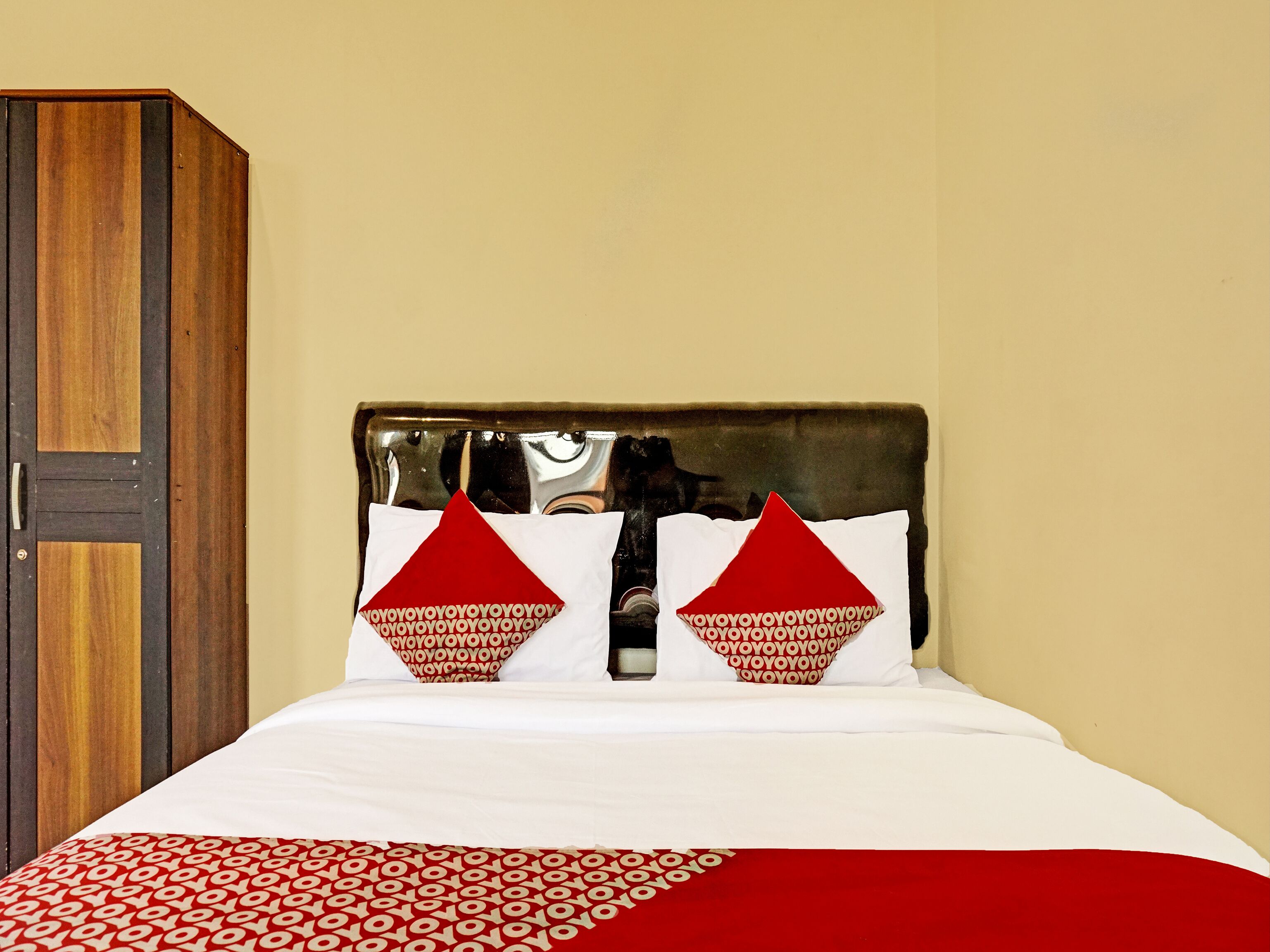 Una cama con ropa de cama blanca y almohadas rojas, un armario de madera y una pared lisa.