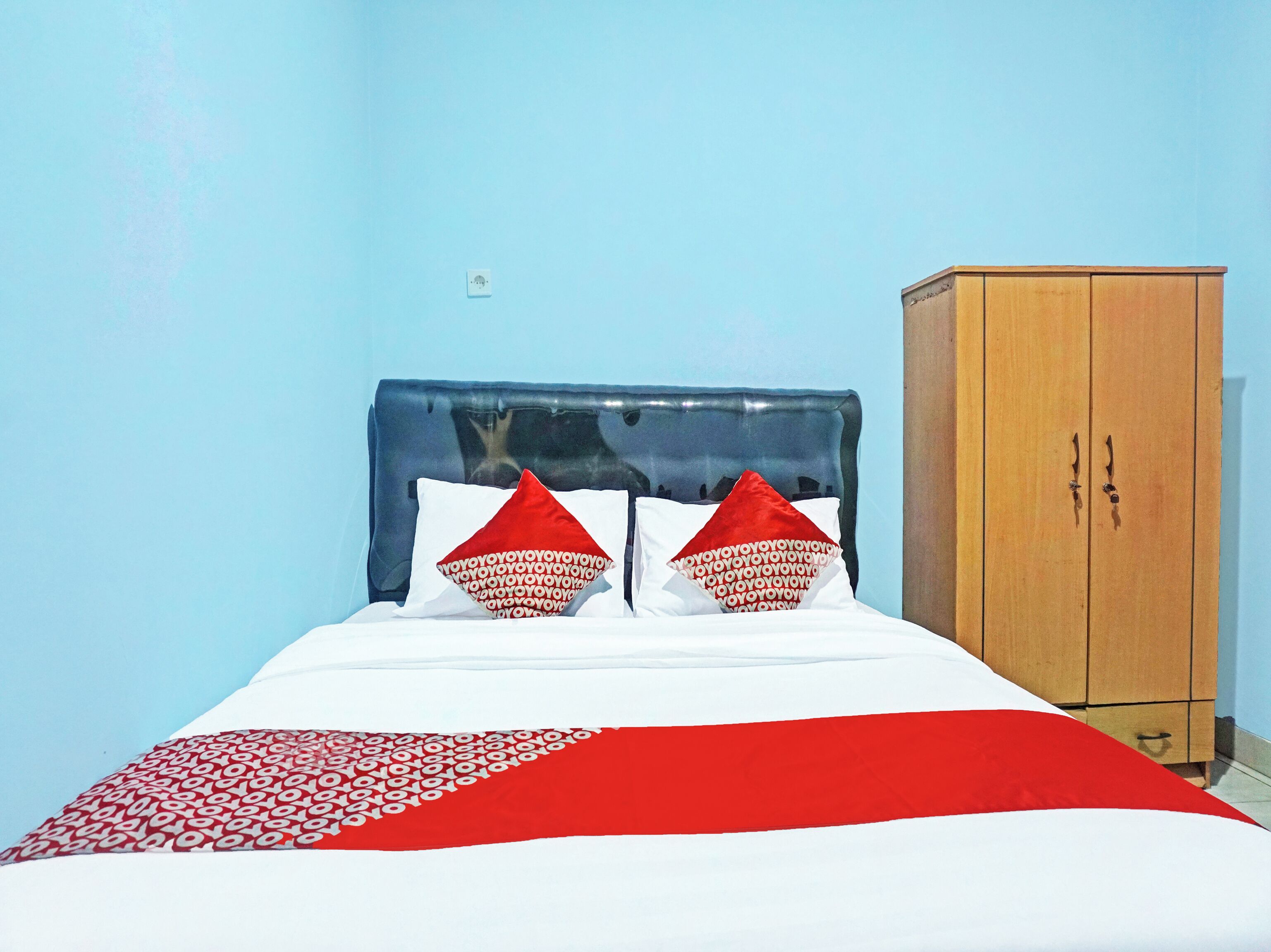 A bed with a red and white bedspread, two red pillows, and a wooden wardrobe against a blue wall.