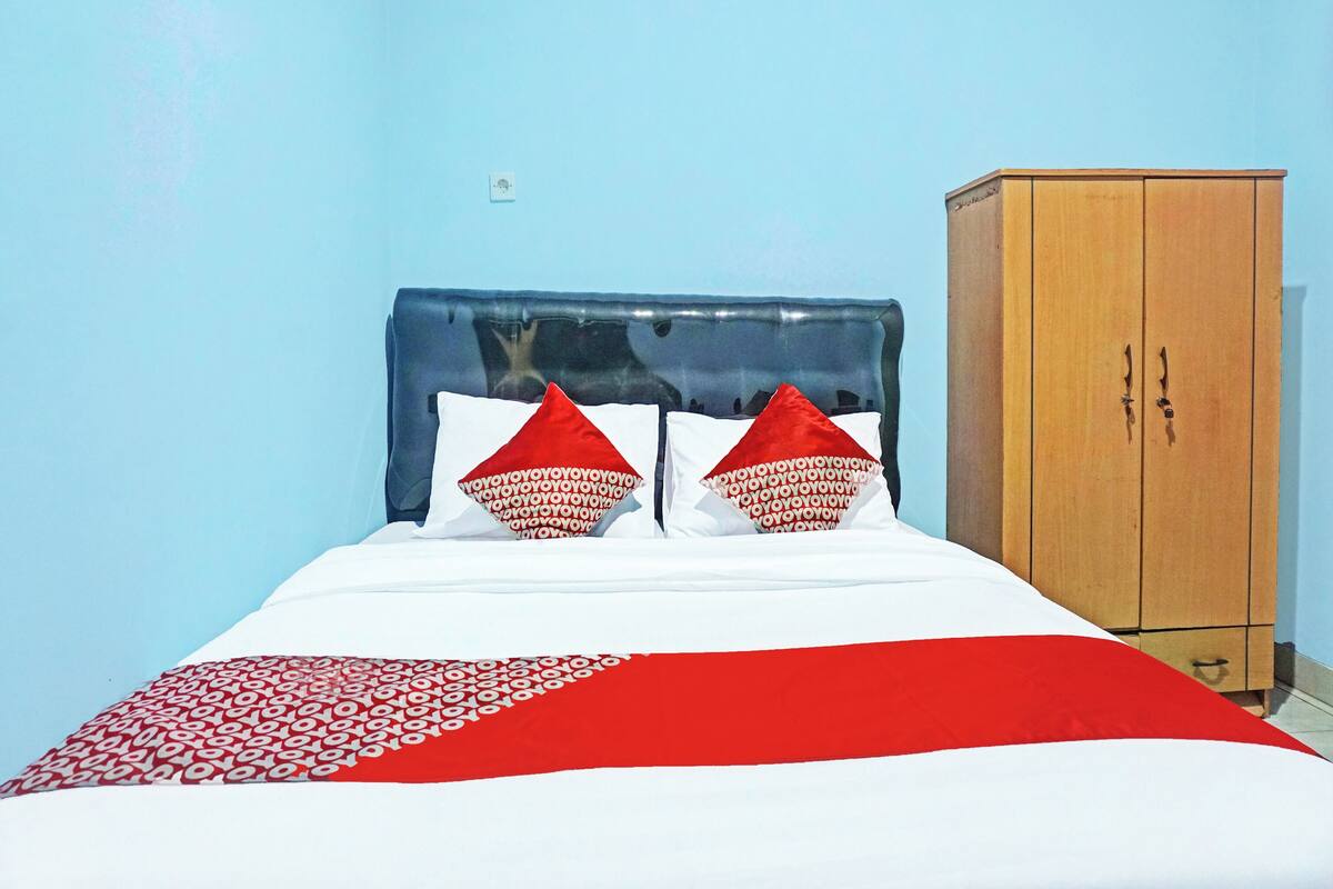 A bed with a blue headboard, two red pillows, and a white and red bedspread. There is a wooden wardrobe to the right.