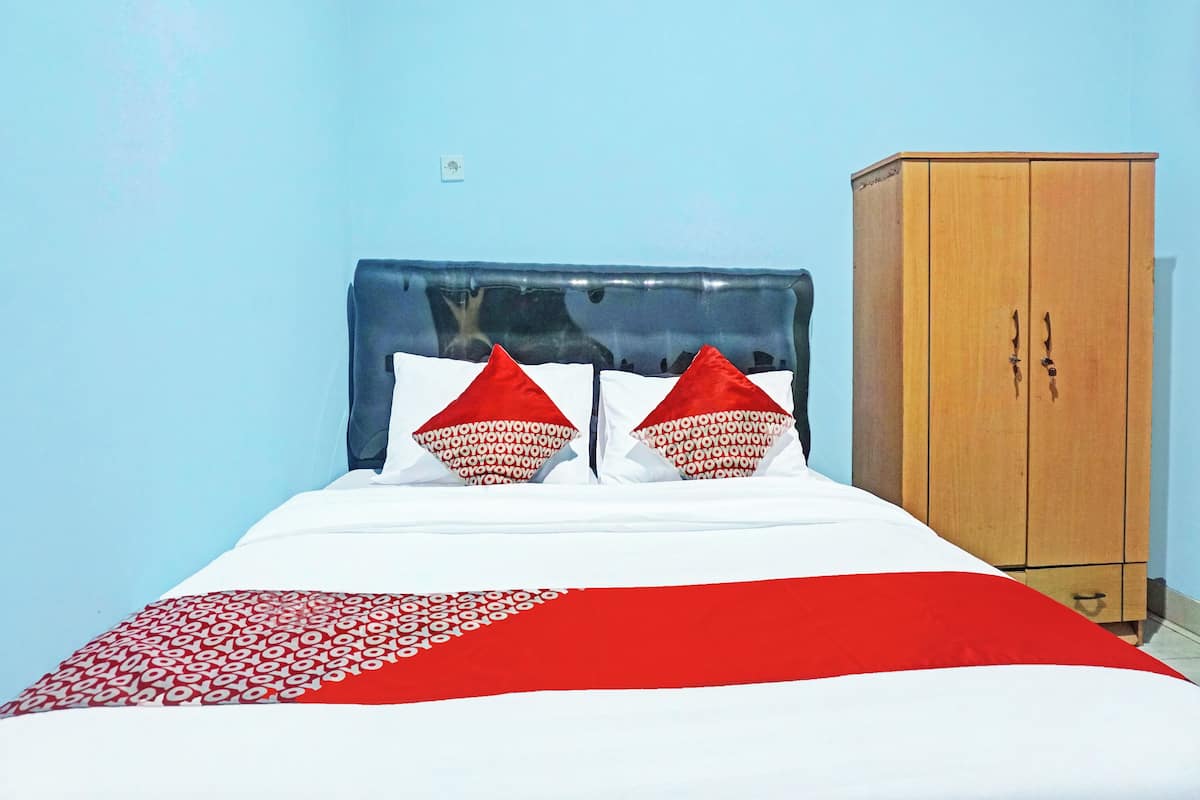 Un lit avec une tête de lit bleue, deux oreillers rouges et une couvre-lit blanche et rouge. Il y a une armoire en bois sur le côté droit.
