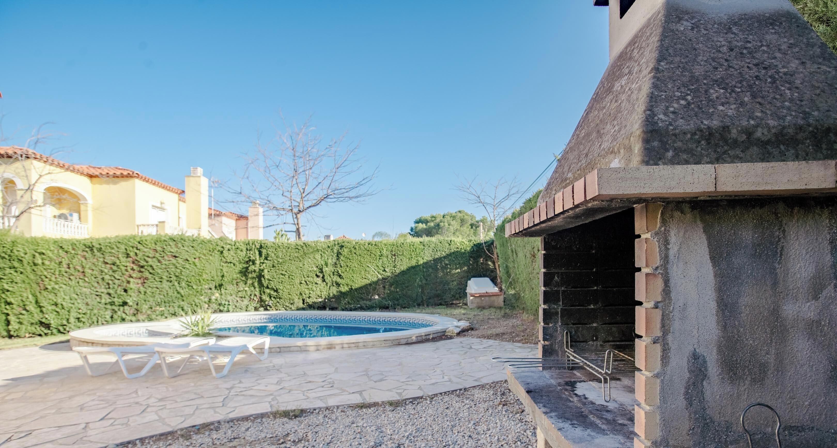 Un espace piscine doté d’une terrasse en pierre, d’un barbecue et d’une haie.