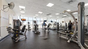 Une salle de sport équipée de divers appareils d’exercice, dotée de grandes fenêtres et d’un ventilateur de plafond.