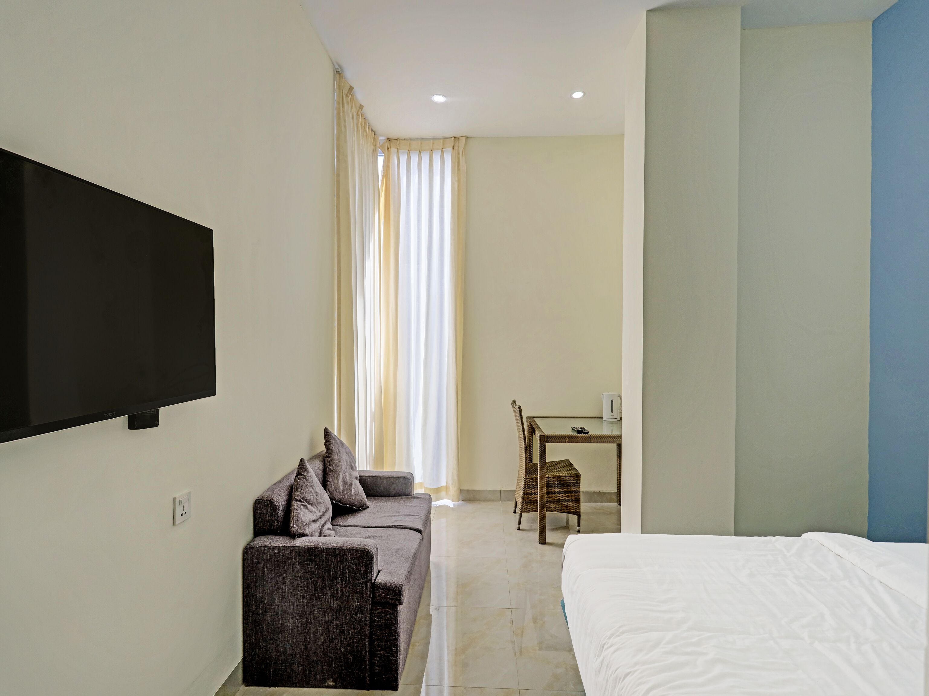 A hotel room with a bed, a sofa, a TV, a table with a chair, and a window with curtains.