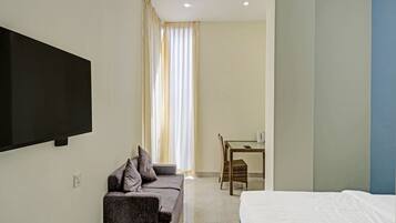 A hotel room with a bed, a sofa, a TV, a table with a chair, and a window with curtains.
