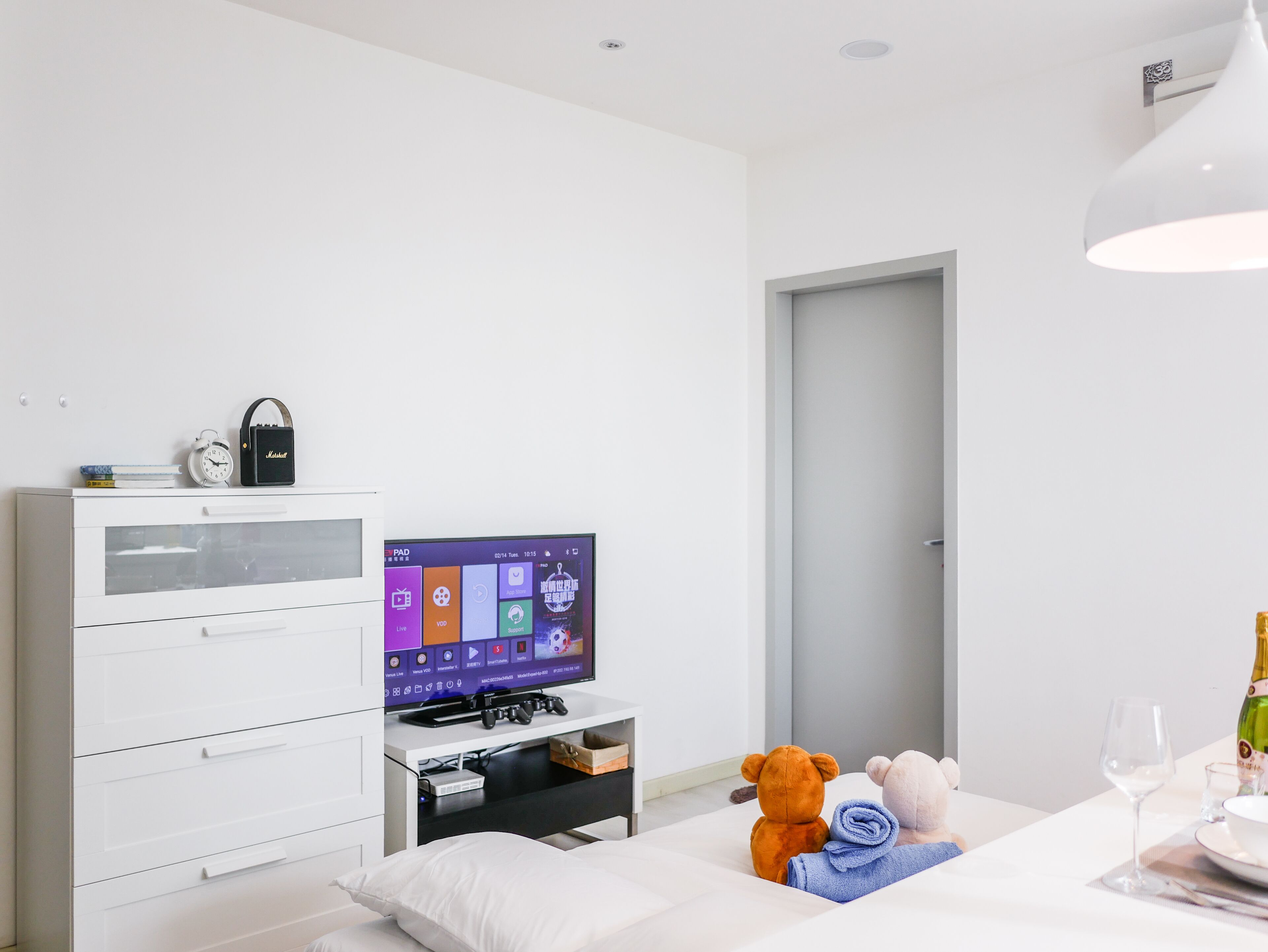 Une chambre d’hôtel moderne avec un lit blanc, un téléviseur posé sur un meuble, un ours en peluche et une bouteille de champagne.