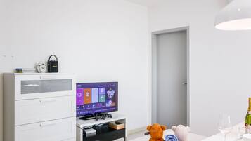 Une chambre d’hôtel moderne avec un lit blanc, un téléviseur posé sur un meuble, un ours en peluche et une bouteille de champagne.