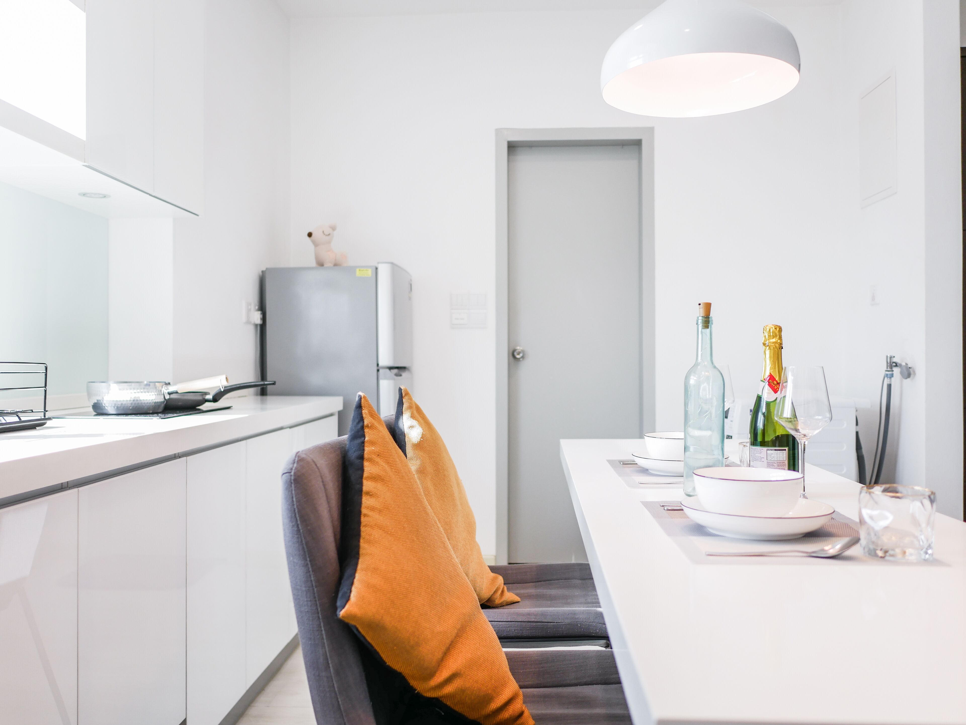 Une cuisine moderne avec des meubles blancs, un réfrigérateur gris et un coin repas avec une table blanche mise pour deux personnes.