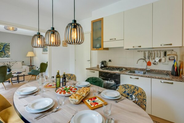 A modern kitchen with white cabinets, a stainless steel sink, and hanging utensils. A dining table is set with plates, glasses, and a bread basket. - Porto Letizia Mimosa 1, Porlezza, Italy (Porlezza)