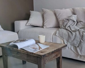 Une pièce avec une table basse en bois sur laquelle reposent un magazine et une tasse, un canapé beige recouvert d’une couverture et un mur uni.