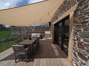 Une terrasse en bois avec une table et des chaises, un mur en pierre et une porte vitrée donnant sur un espace intérieur.