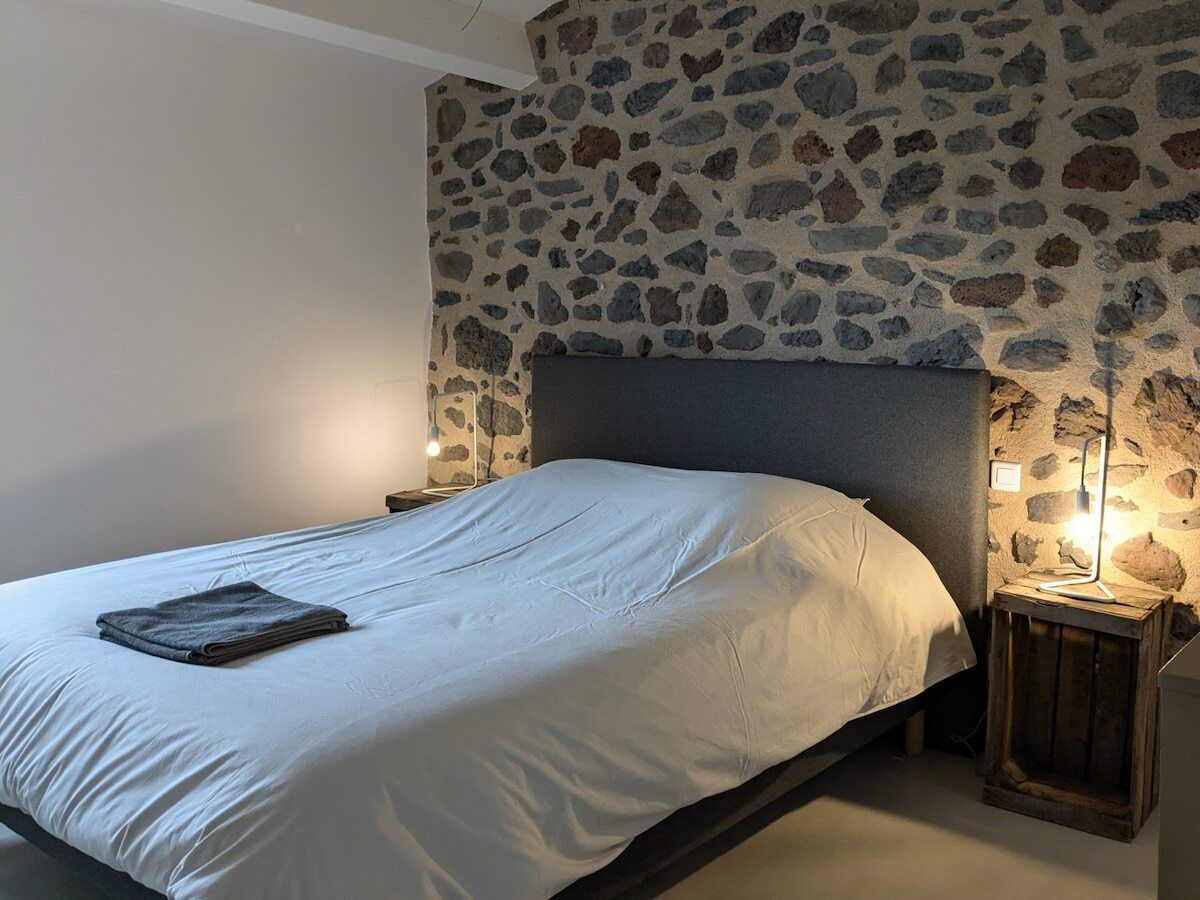 A bed with a white mattress and a folded blanket, a stone wall, and two bedside lamps.