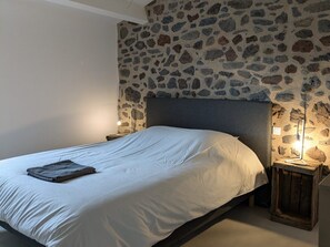 Un lit avec un matelas blanc et une couverture pliée, un mur en pierre et deux lampes de chevet.