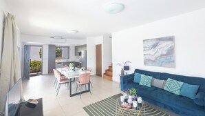 Una sala de estar moderna con un sofá azul, una mesa de comedor y sillas. Se observa una cocina al fondo, una escalera y un ventanal grande.