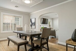 Uma sala de jantar com uma mesa de madeira escura, cadeiras acolchoadas e um banco. Há uma janela grande com persianas, uma televisão de tela plana e um quadro na parede.