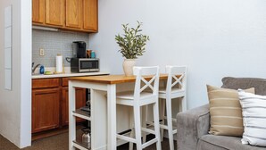 Cocina compacta con armarios de madera, microondas y cafetera. Hay una mesita con dos sillas blancas y una planta en maceta. Se ve un sofá gris con cojines a rayas al fondo.