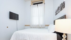 Un dormitorio con cama, televisor montado en la pared, ventana con cortinas blancas y lámpara en la mesita de noche.