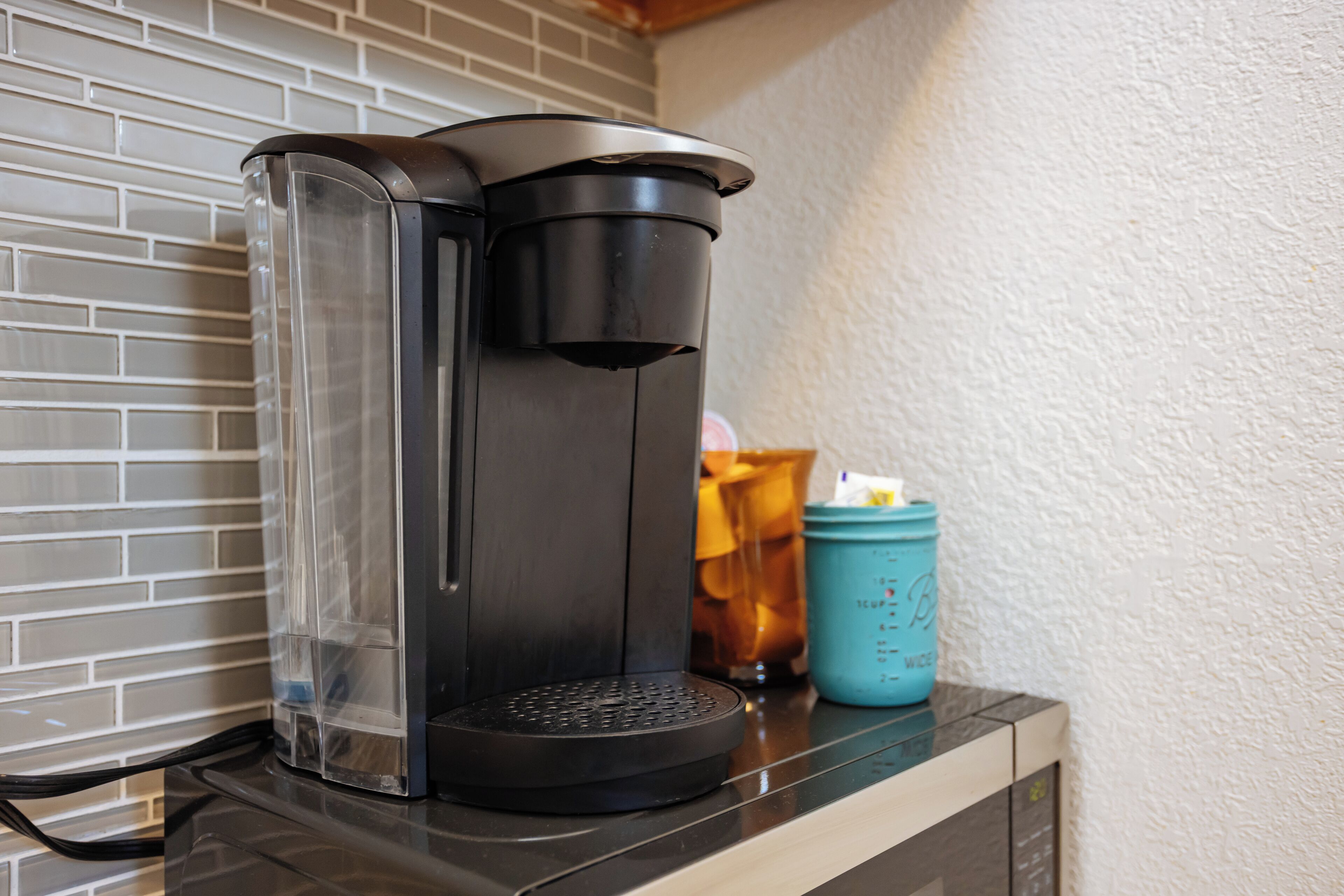 Una cafetera con depósito de agua transparente y bandeja de goteo negra, sobre una encimera junto a una taza azul.